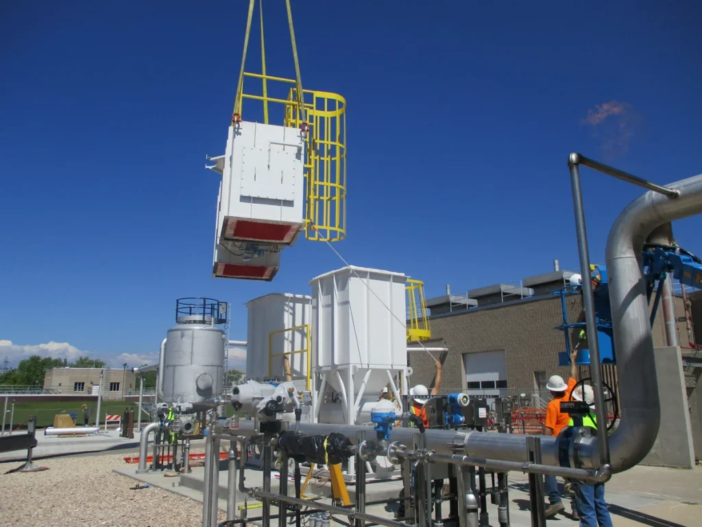 Workers guiding crane-lifted equipment section into position among piping at treatment facility
