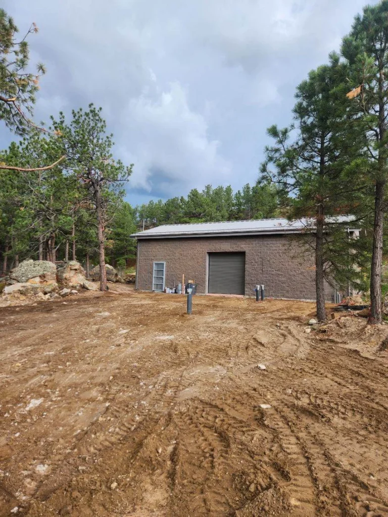 Newly constructed pump station building with garage door surrounded by pine trees and cleared ground