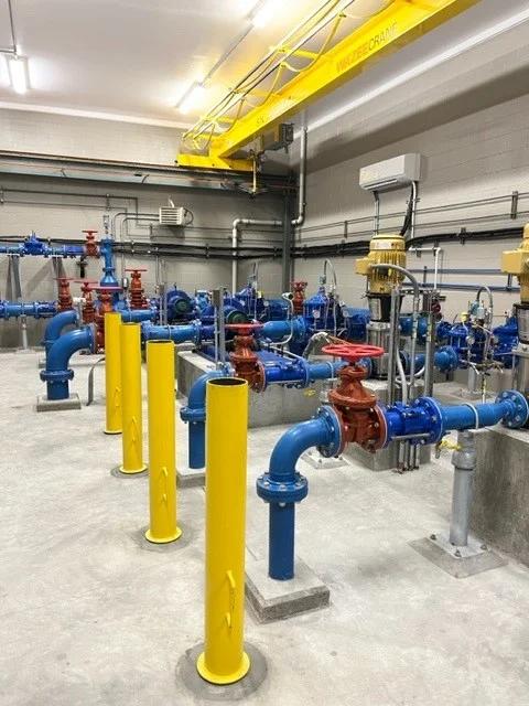 Pump station interior showing blue piping, red valves, yellow bollards, and overhead bridge crane