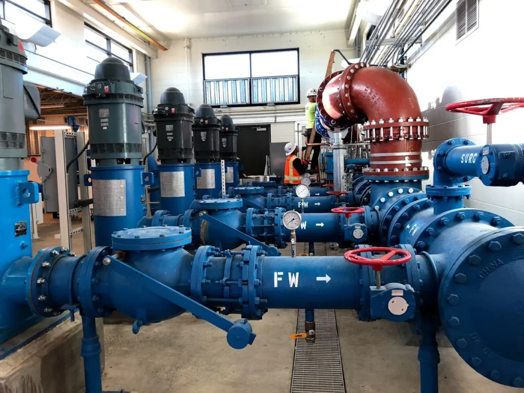 Interior of well house showing blue finished water piping system with pumps, valves, and gauges