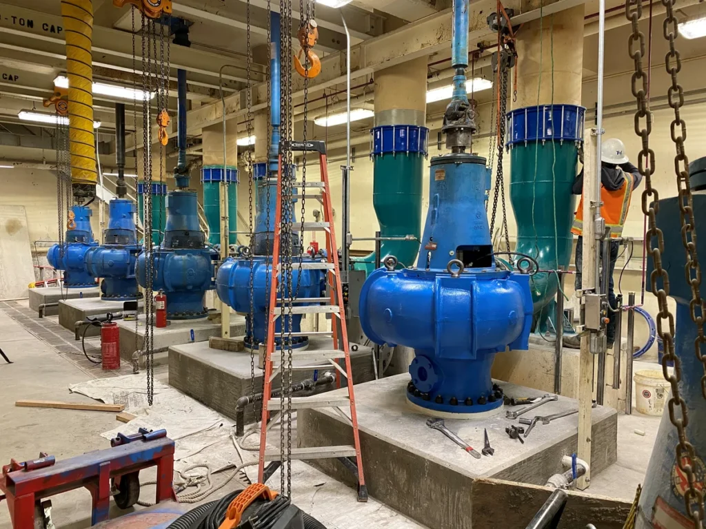 Row of large blue vertical turbine pumps being installed with chains and overhead crane