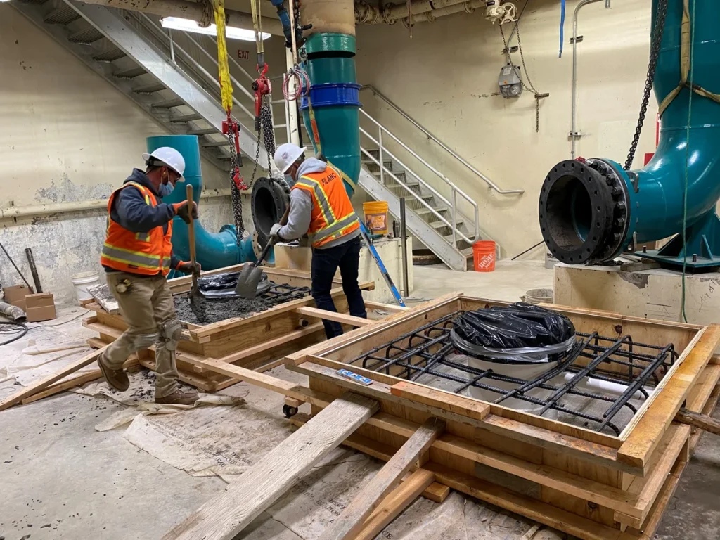 Construction workers pouring concrete around new piping in treatment facility pump room